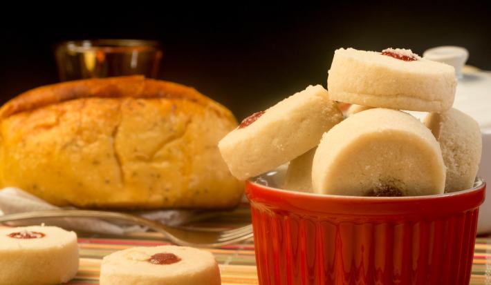 Fotografia de biscoitinhos amanteigados recheados com docinho de Leite MOÇA e NESQUIK de morango dentro de um recipiente de vidro branco e vermelho. Ao lado tem dois biscoitinhos jogados e no fundo tem um pão.