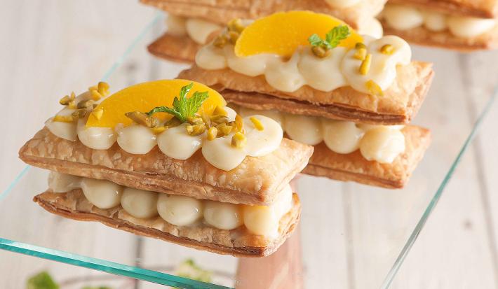 Fotografia em tons de amarelo em uma bancada de madeira com um suporte para bolo de vidro e quatro mil folhas com recheio de creme e pêssegos.