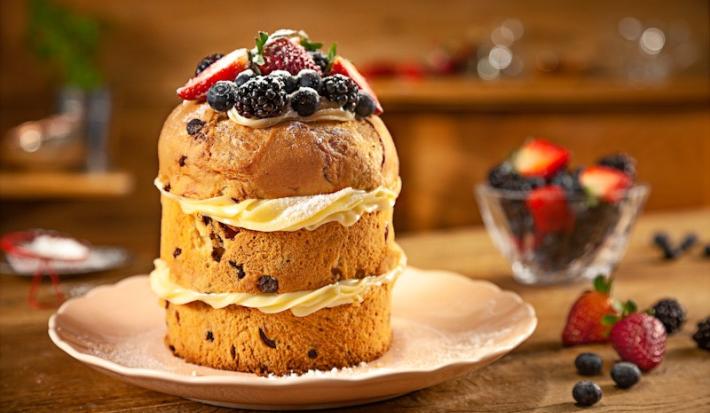 Fotografia em tons de marrom de uma bancada marrom com um prato rosa, sobre ele um panettone recheado com frutas vermelhas no topo. Ao lado um recipiente de vidro com frutas vermelhas.