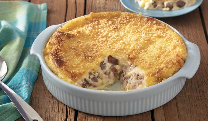 Foto em tons de amarelo da receita de escondidinho de carne de sol servida em uma porção grande em uma forma branca sobre uma mesa de madeira com um pano verde e azul ao lado