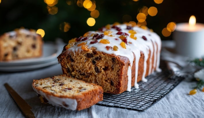 Fotografia de um bolo de frutas, com uma fatia cortada em destaque. O bolo é decorado com glacê branco e frutas secas coloridas. A cena é iluminada por uma vela, transmitindo um clima natalino.