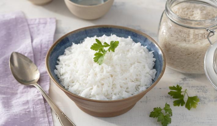 Fotografia em tons de lilás em uma bancada de madeira clara, um pano lilás e um pote marrom redondo fundo com o arroz basmati dentro dele. Ao lado, um pote hermético com arroz cru e uma colher de servir.