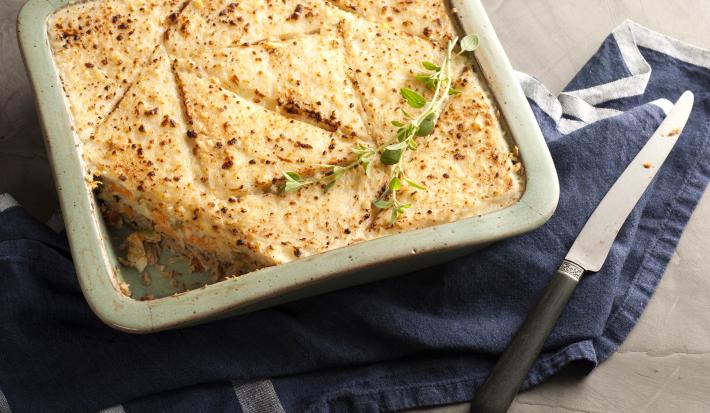 Fotografia em tons de dourado e azul, assadeira com a torta de pão de forma gratinada com queijo e folhas verdes, sobre guardanapo azul com listras brancas, faca ao lado, tudo sobre toalha cinza.