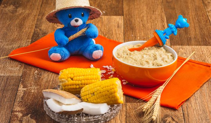 Fotografia em tons de laranja e azul de uma bancada de madeira com um paninho laranja e um potinho com uma papinha dentro. A frente uma espiga de milho ao meio, pedaços de coco e atrás um ursinho de pelúcia azul com um chapéu de palha.