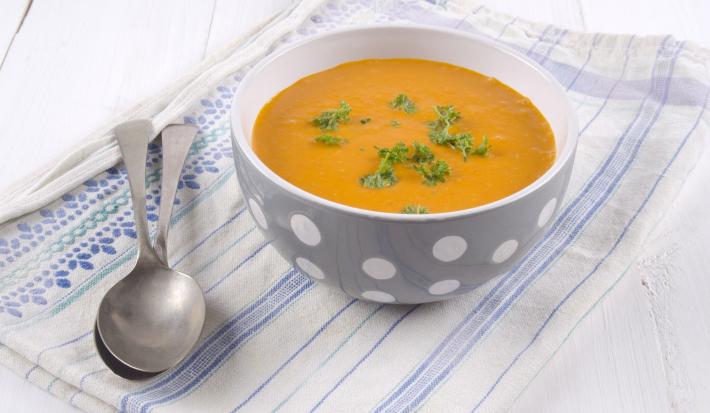 fotografia em tons de branco, laranja e azul de uma bancada branca vista de cima. Contém um pano branco com listras azuis, por cima um recipiente redondo cinza com bolinhas brancas e dentro a sopa. Ao lado do recipiente duas colheres.