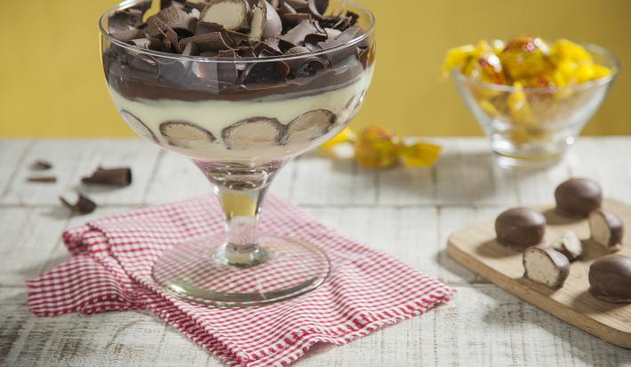 Fotografia em tons de amarelo e chocolate, ao centro taça com creme, bombons cortados ao meio e raspas de chocolate sobre guardanapo xadrez branco e vermelho, ao lado tábua com bombons, ao fundo potinho com bombons, tudo em bancada de madeira em pátina.