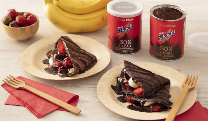 Foto em tons de vermelho da receita de crepioca doce feita com Nescau extra cacau servida em duas porções em pratos de madeira sobre panos vermelhos. Ao fundo, morangos e bananas de decoração, além das latas de nescau e garfos de madeira