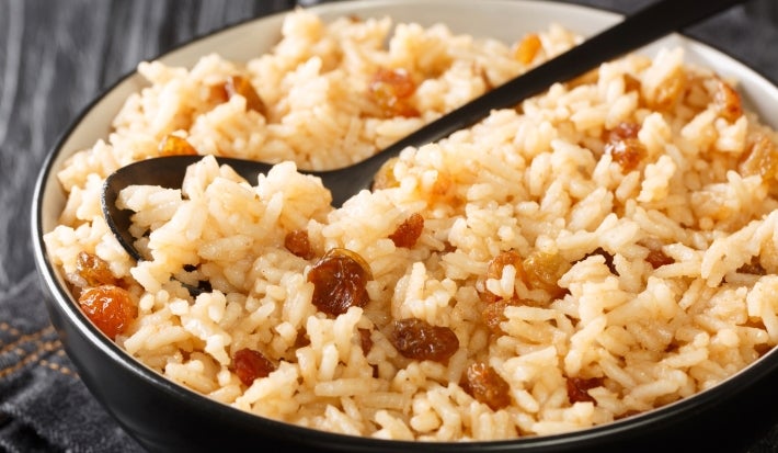 Em cima de uma mesa, tigela com uma colher preta com arroz soltinho com uvas-passas brancas.