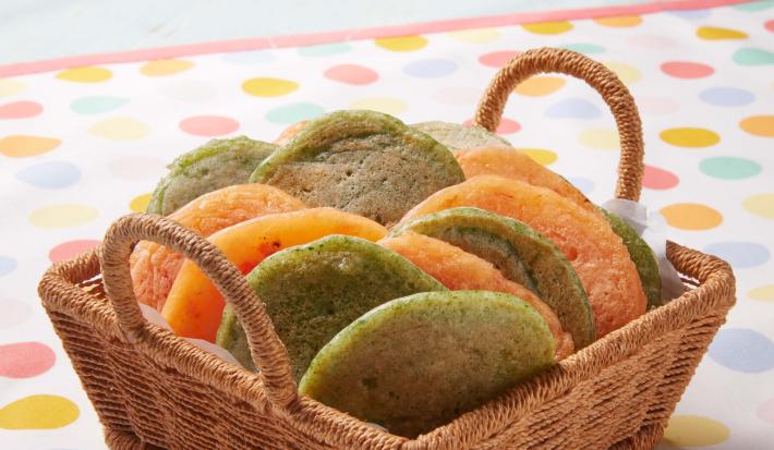 Fotografia em tons de verde e laranja em uma bancada de madeira, uma toalha branca com bolinhas coloridas, uma cesta de vime marrom clara com pães de queijo achatados nas cores verde e amarelo.