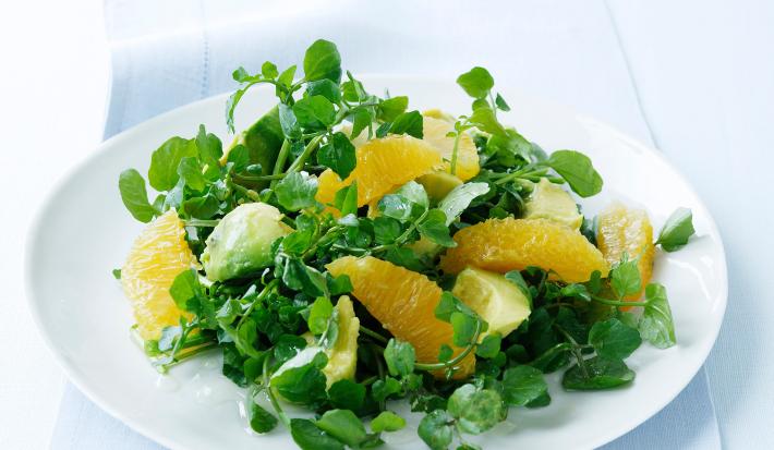 Fotografia em tom claro, um prato branco com uma salada de agrião e pedaços de tangerina, sobre uma toalha de mesa azul em tom claro.