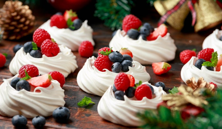 Imagem de mini pavlovas de iogurte decoradas com frutas vermelhas sobre uma mesa de madeira e decoração de Natal.