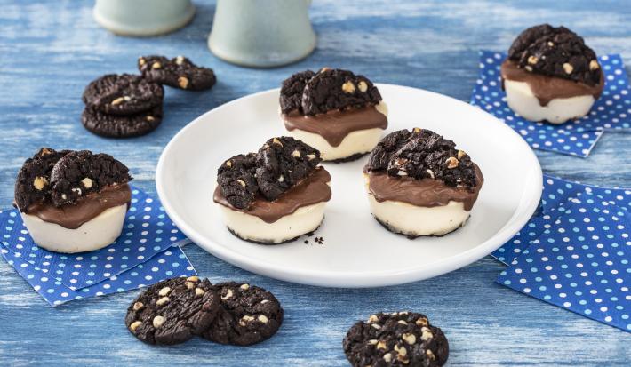 Foto de uma bancada de madeira azulada, com guardanapos azuis de bolinhas brancas espalhados e, ao centro, um prato branco com três mini cheesecakes decoradas com Cookies Negresco. Há mais cookies Negresco espalhados pela bancada