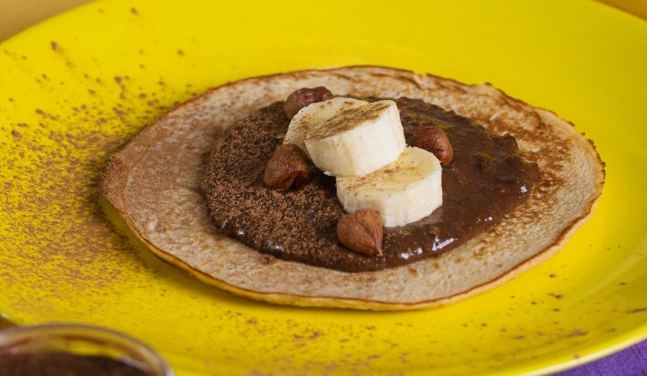 Fotografia de um prato amarelo com uma panqueca de banana com bananas por cima, creme de avelã e três aveãs inteiras. .