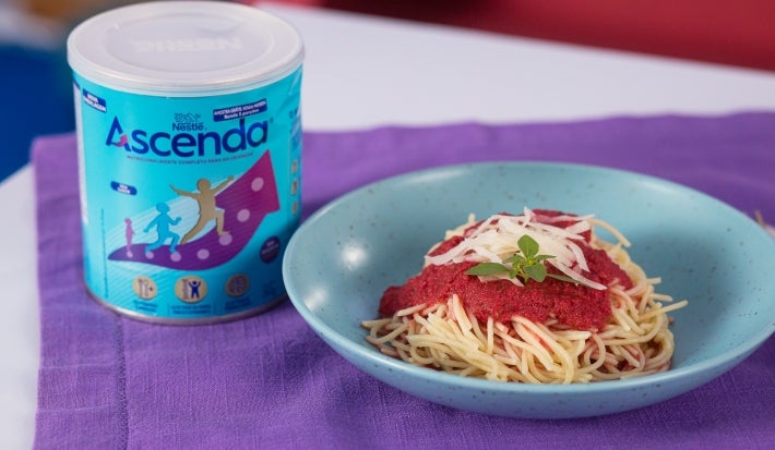 Fotografia de um prato fundo azul, pequeno, sobre um pano roxo, ao lado de uma lata de Ascenda. Dentro do prato tem macarrão tradicional com o molho de tomate e ascenda.