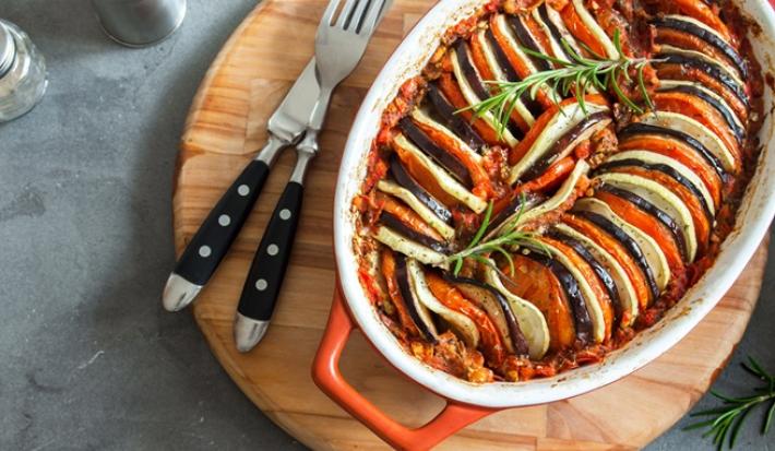 Fotografia em tons de vermelho com uma tabua de madeira ao centro. Em cima da tabua existe uma receita de ratatouille feita com berinjela, abobrinha, tomate e alecrim