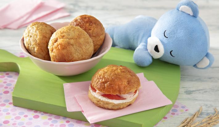 Foto da receita de Pão de Leite. Observa-se um potinho rosa com três pãezinhos e, ao lado direito, um pão com recheio de tomate, requeijão e peito de peru, sobre uma tábua verde.