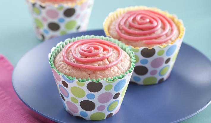 Fotografia em tons de azul e rosa em uma mesa de madeira azul, um pano rosa, um prato azul redondo com dois cupcakes de morango com aveia em forminhas coloridas.