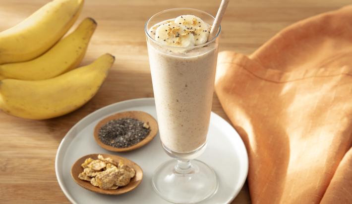 Foto da receita de Smoothie de Banana com Nesfit Cereal. Observa-se um copo alto com o smoothie dentro e finalizado com NESFIT Cereal, banana e chia. Ao lado esquerdo, potinhos com NESFIT cereal, chia e um cacho de bananas decoram a foto.