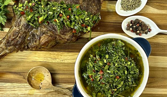 Foto da receita de Chimichurri. Observa-se um carré de cordeiro com o molho por cima sobre uma tábua de madeira.