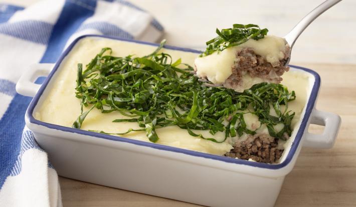 Fotografia em tons de azul em uma bancada e madeira, um pano xadrez azul e ao centro um recipiente refratário branco com detalhes em azul e o escondidinho de carne e batata e couve por cima para decorar.