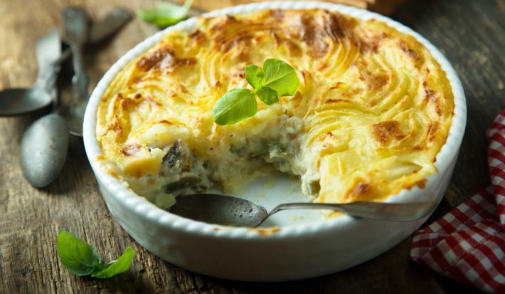 Foto em tons de amarelo da receita de torta de bacalhau servida em uma forma de porcelana branca com uma fatia cortada separada da torta e uma colher prateada dentro
