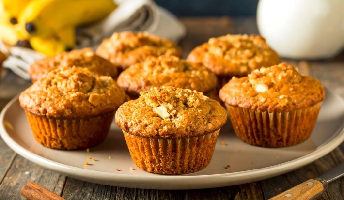 A imagem mostra vários muffins de banana com uma cobertura crocante, dispostos em um prato claro sobre uma mesa rústica de madeira. Os muffins têm uma cor dourada e textura levemente crocante por fora, com aparência macia por dentro.