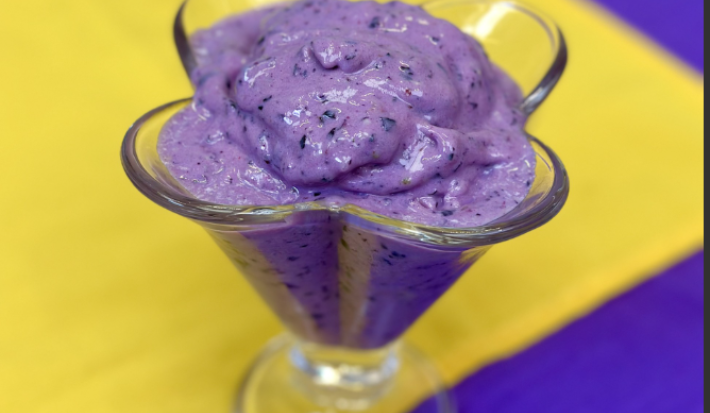 sobre um fundo azul, amarelo e branco, dentro uma taça de vidro transparente está um sorvete cremoso em tons de roxo com pedacinhos de uva.