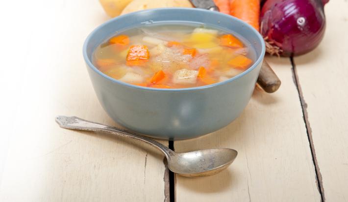 Fotografia de uma sopa de batata, cenoura e chuchu, dentro de um recipiente de vidro na cor azul. Na frente, uma colher de sopa, e ao fundo, cebolas roxa, batatas e cenoura, sobre uma mesa de madeira.