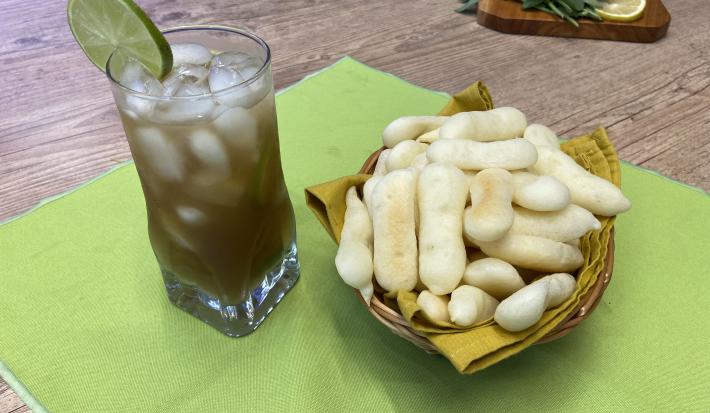 Fotografia em tons de verde em uma bancada de madeira, um pano verde, uma cesta de vinil com guardanapo amarelo com os biscoitos de polvilho em cima, ao lado um copo de vidro alto com o chá verde, cubos de gelo e uma rodela de limão decorando.