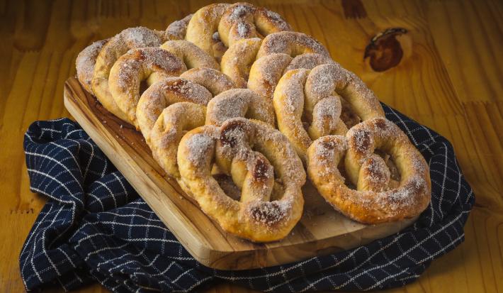 Foto aproximada de uma bancada de madeira sobre a qual está um pano azul quadriculado e nele há uma tábua de madeira com alguns pretzels decorados com açúcar e canela