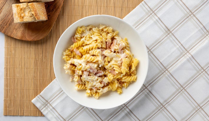 Prato branco com macarrão parafuso com bacon e molho cremoso sobre uma toalha xadrez e uma tábua com fatias de pão.