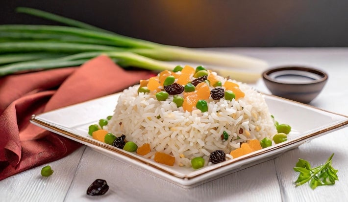Em um prato branco, porção de arroz branco com uvas-passas, ervilha e cenouras picadas em cubos.