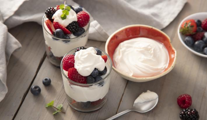 Fotografia em tons de vermelho em uma bancada de madeira clara, um pano cinza, um potinho com o mascarpone, um potinho com frutas vermelhas. Ao centro, dois copos de vidro com frutas vermelhas cobertas por marcarpone.