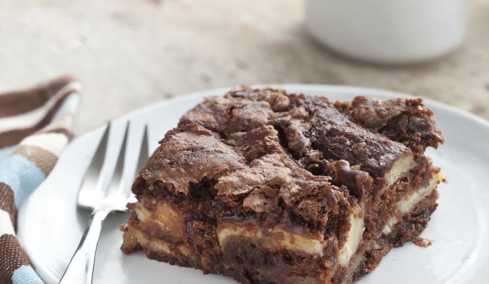 Fotografia em tons de azul e marrom em uma bancada de madeira clara, um pano listrado em azul e marrom, um prato branco redondo raso com um pedaço de torta de banana com chocolate.