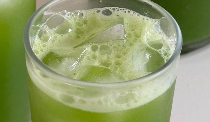 Fotografia de três copos de vidro com suco verde e fiber mais de limão. Os copos estão sobre uma mesa branca, e dentro deles, com o suco, tem cubos de gelo.