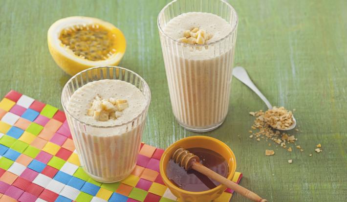 dois copos transparentes com a vitamina dentro ao lado um potinho redondo na cor amarelo com mel dentro e ao fundo uma fatia de maracujá