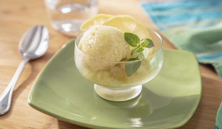 Fotografia em tons de verde em uma bancada de madeira de cor marrom. Ao centro, um prato quadrado verde contendo uma taça com o sorbet. Ao lado, há uma colher e ao fundo um pano azul.