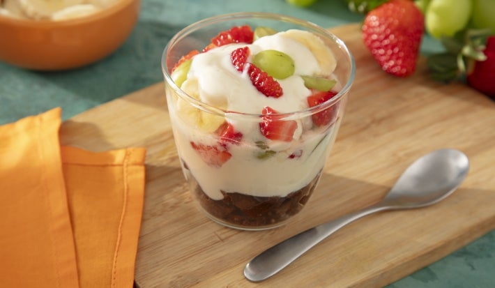 Fotografia mostra uma taça de pavê com camadas visíveis de creme branco, frutas coloridas e pedaços de biscoito integral, formando um visual fresco e apetitoso.