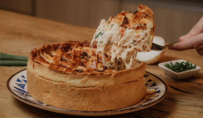 Fotografia em tons de marrom de uma bancada de madeira com um prato raso branco com detalhes azul sobre ele um quiche de frango com um pedaço sendo cortado. Ao fundo um potinho branco quadrado com cebolinha e uma cebola.