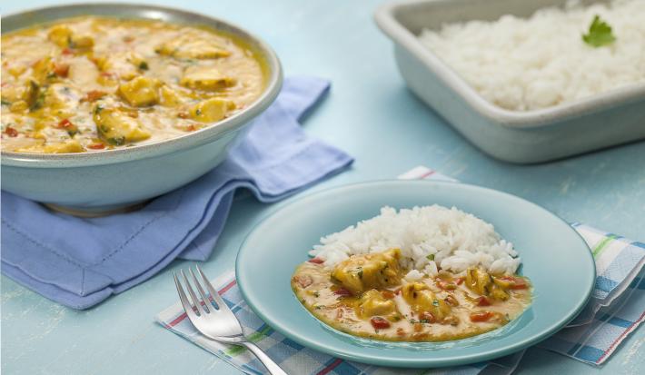 Fotografia em tons de azul e amarelo, ao centro prato azul com frango ensopado e arroz, sobre guardanapo azul e branco com um garfo, à direita travessa com arroz, à esquerda travessa com frango ensopado e guardanapo azul, tudo sobre bancada na cor azul.