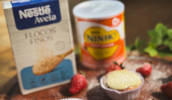 Fotograifa em tons claros da receita sob o sol de três muffins em forminha de papel sobre uma tábua grande e redonda de madeira. Tem ao lado dos muffins alguns morangos inteiros com leite ninho polvilhado.