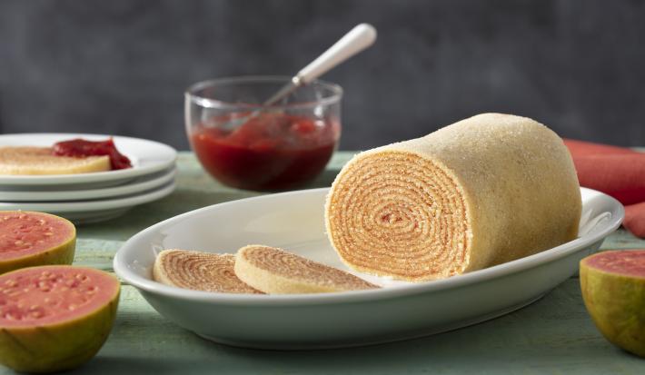Imagem de um prato oval com o bolo de rolo e duas fatias cortadas. Ao fundo, uma pote de goiabada e goiabas ao lado.