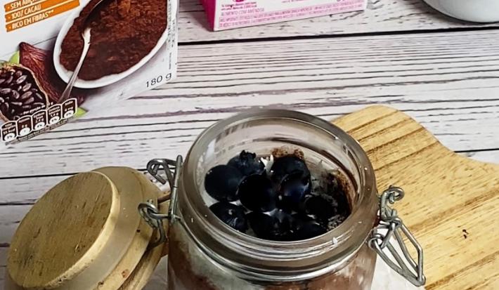 Foto da receita de pudim de chia servida em um pote hermético de vidro sobre uma tábua de madeira em cima de uma mesa de madeira branca. Ao fundo há embalagens de nature's heart cacau em pó e bebida vegetal de amêndoa