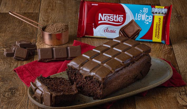 Foto da receita de Chocolate Bar Cake, sobre uma bancada de madeira, em um prato em tom marrom. Na bancada há uma barra de chocolate ao leite, um copo de medida de cobre com cacau em pó e alguns pedaços de chocolate