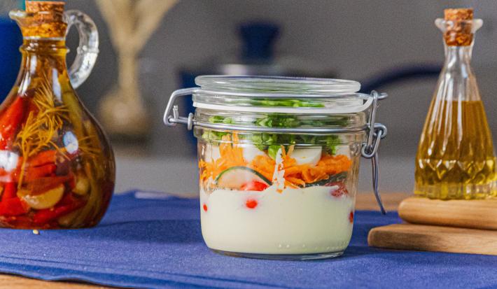Fotografia em tons de azul com um pote de vidro ao centro. Dentro do pote existe uma receita de salada rúcula com cenoura, pepino, tomate cereja com molho caesar