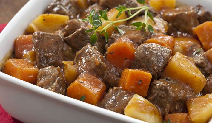Fotografia em tons de marrom com um paninho vermelho e um recipiente branco retangular com picadinho de carne com batatas e cenoura