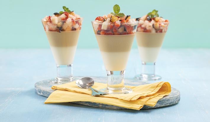 Foto em tons de azul da receita de pudim de coco com calda de ameixa servida em 3 taças de vidros com pedaços das frutas por cima, as taças sobre uma tábua de vidro com um pano amarelo e uma colher prateada para decoração.