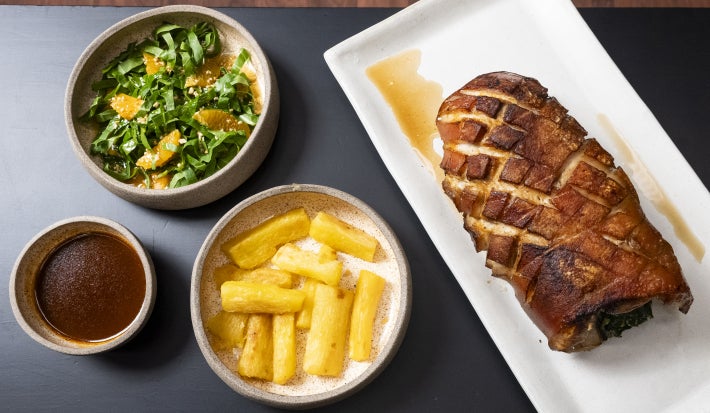 Pernil suíno assado com mandioca dourada, salada de folhas com laranja e molho servido à parte, apresentado em travessas.