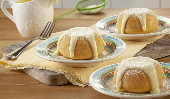 Fotografia em tons de bege com três porções de bolinhos com cobertura em pratinhos coloridos, sobre guardanapo amarelo e tábua de cozinha com um garfo ao lado, pote de cobertura e peneirinha verde ao fundo em bancada de madeira.