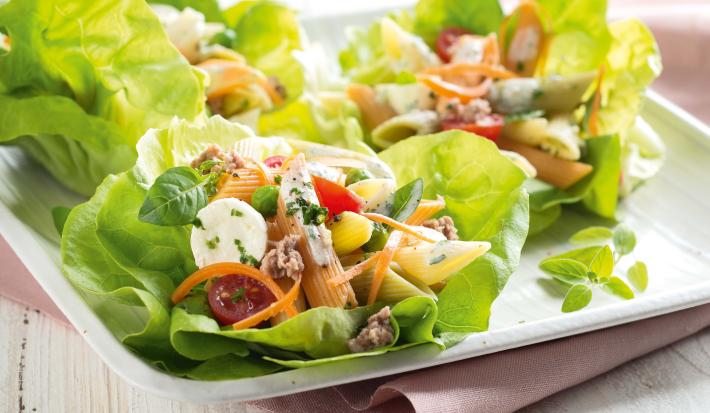 Fotografia em tons de branco e verde, ao centro salada de folhas verdes com macarrão, legumes e mozarela, sobre guardanapo rosa e bancada de madeira em tons de branco.